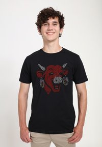 Schwarzes T-Shirt mit einem stilisierten roten Kuhkopf mit weißen Akzenten und bunten Ohrringen, präsentiert an einem Model vor einem neutralen Hintergrund.