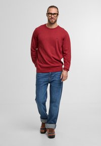 Roter Pullover mit Rundhalsausschnitt, langen Ärmeln und gerippten Bündchen, kombiniert mit blauen Jeans und braunen Wanderschuhen. Modell steht vor einem schlichten Hintergrund.