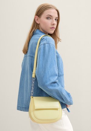 Borsa a spalla in pelle vegan gialla con chiusura a pattina, dotata di una tracolla con catena e dalla forma arrotondata. Indossata sopra una giacca di jeans.