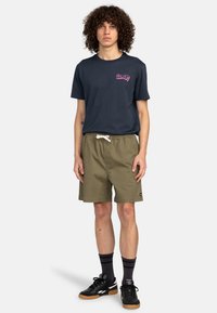 Marineblaues T-Shirt mit pinkem Logo, olivgrüne Shorts mit Kordelzug, schwarze Sneaker und gestreifte schwarze Socken. Ein lässiger, bequemer Schnitt.