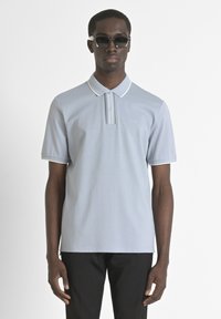 Polo bleu clair avec des bordures blanches et une patte de boutonnage à deux boutons, fabriqué à partir d'un matériau lisse. Porté avec un pantalon noir et des lunettes de soleil.