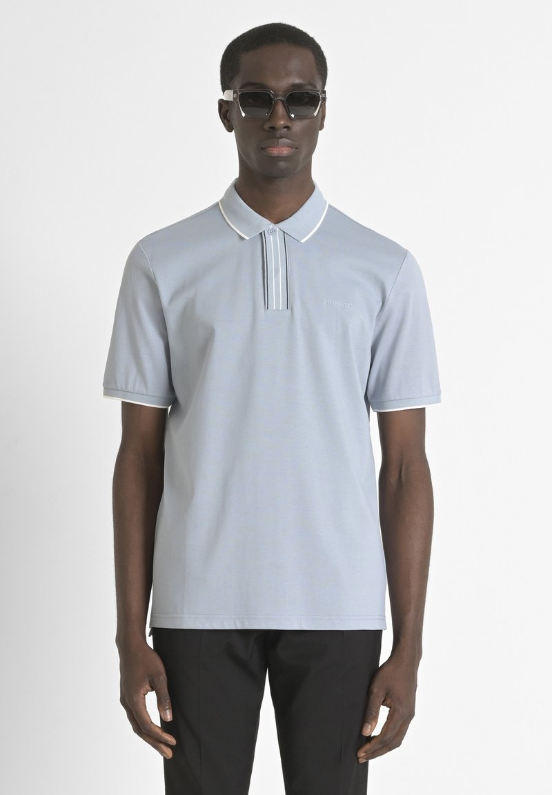 Polo bleu clair avec des bordures blanches et une patte de boutonnage à deux boutons, fabriqué à partir d'un matériau lisse. Porté avec un pantalon noir et des lunettes de soleil.