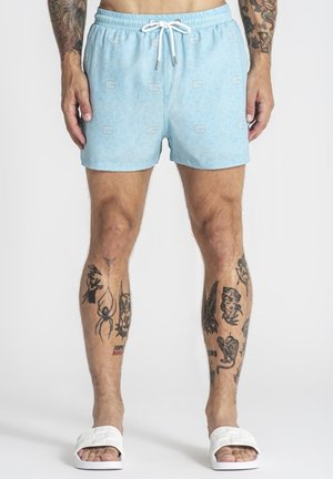 Homme portant un short de bain bleu clair à motifs avec un cordon blanc, des sandales blanches et plusieurs tatouages sur les jambes et les bras.