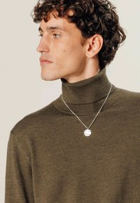 Pull à col roulé vert olive en texture côtelée, arborant un collier argenté avec un pendentif circulaire, porté par un mannequin masculin.