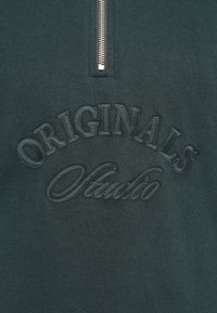 Donkergroene sweatshirt met een halve rits en geborduurde tekst "ORIGINALS Studio" met een gestructureerde afwerking.