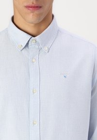 Világoskék, gombos ing, függőleges csíkos mintával. Jellemzői a hegyes gallér, fehér gombok és az emelt logó a mellkason.