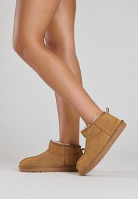 Bottines en suède marron avec une doublure en fourrure douce, semelle en caoutchouc plate et tirette à l'arrière, posées sur un fond clair.