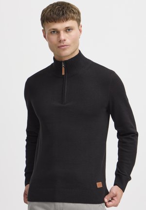 Schwarzer Pullover mit hohem Kragen, ausgestattet mit einem halben Reißverschluss und einem gerippten Saum. Hergestellt aus einem glatten, strukturierten Material mit einem Lederlogo-Patch.