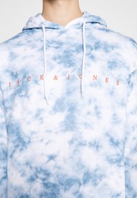 Niebieska i biała bluzka z kapturem w technice tie-dye, z troczkami w kapturze i napisem "JACK & JONES" w pomarańczowym kolorze na piersi. Miękka tekstura materiału.
