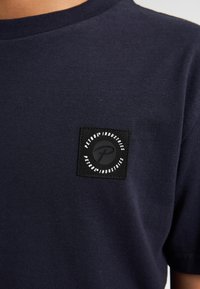 Donkerblauw katoenen T-shirt met een ronde hals en korte mouwen, voorzien van een zwart vierkant stukje met witte tekst en logo op de borst.