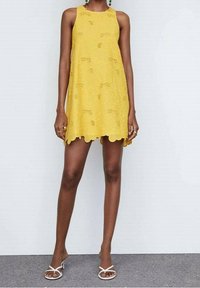 Żółta sukienka bez rękawów z kwiatowym koronkowym wzorem, falowanym dołem i luźnym krojem; zestawiona z białymi sandałami na obcasie.
