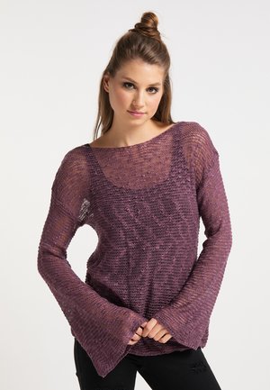 Jonge vrouw met een lichte huid en bruin haar in een knot, draagt een losse paarse gebreide trui over een zwart hemdje, staand met de handen in elkaar gevouwen.