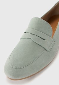 Mocassin en suède vert avec une couture et un onglet décoratif sur le dessus. Possède une texture lisse et un design à bout arrondi.