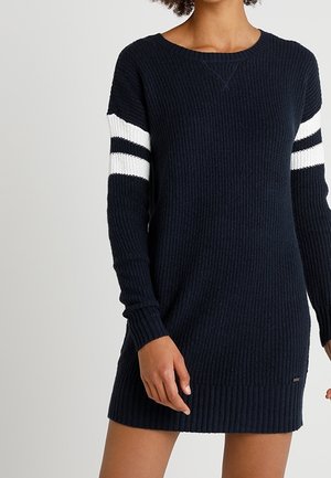 Frau trägt ein marineblaues gestricktes Pulloverkleid mit weißen und schwarzen Streifen an den oberen Ärmeln, steht vor einem einfarbigen Hintergrund.