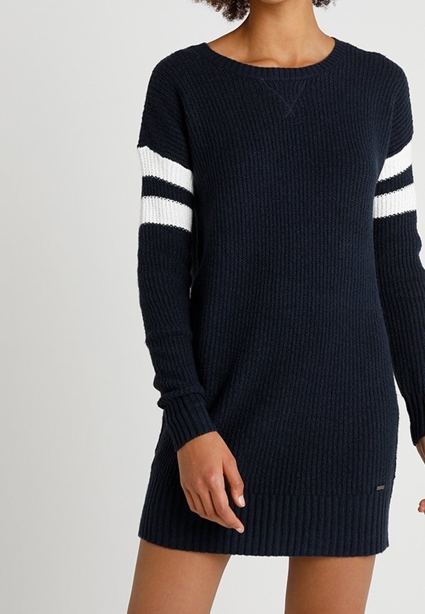 Femme portant une robe-pull en maille bleu marine avec des rayures blanches et noires sur le haut des manches, debout devant un fond uni.