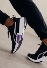 Baskets de sport noires avec des accents violets et blancs, en tissu en maille, à bout arrondi et semelle en caoutchouc, avec un design lacé et des motifs texturés.