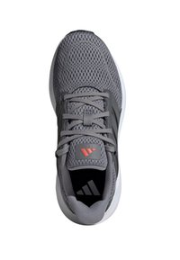 Zapatilla deportiva gris con parte superior de malla, cordones texturizados y acentos negros. Presenta un logo prominente en la lengüeta y una suela blanca.