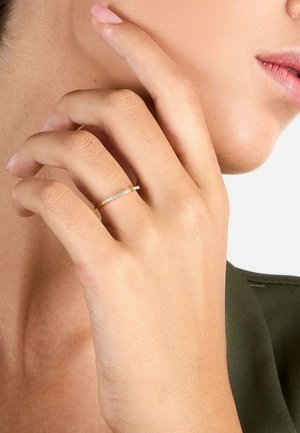 Hand with pale pink nail polish wearing a thin gold ring with small diamonds on the ring finger near a woman’s jawline.