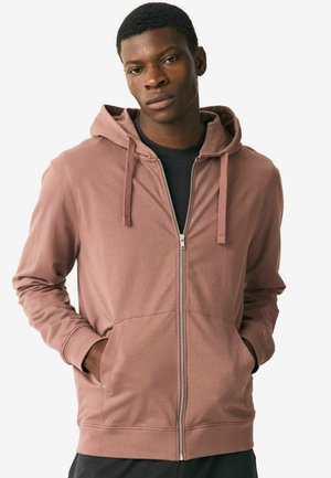 Sudadera con cremallera de color rosa claro, con grandes bolsillos delanteros, cordones y mangas largas, llevada sobre una camiseta negra. Textura de tela suave y lisa.