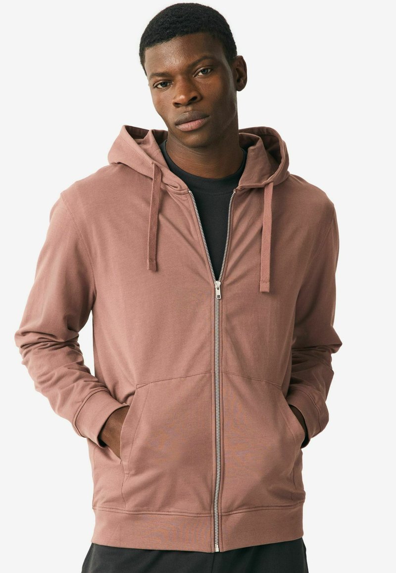 Light pink zip-up hoodie with large front pockets, drawstrings, and long sleeves, worn over a black shirt. Smooth, soft fabric texture.