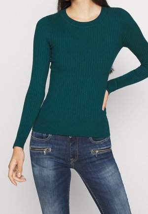 Femme portant un pull vert ajusté à manches longues côtelé et un jean bleu foncé avec poches zippées à l'avant, posant avec une main sur la hanche.