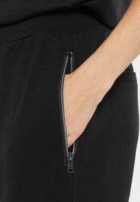 Schwarze Hose mit strukturiertem Finish, ausgestattet mit einer Reißverschlusstasche an der Seite. Der Reißverschluss ist silber mit einem Zugband, und eine Hand befindet sich in der Tasche.