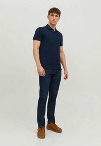 Μπλε κοντομάνικο πόλο μπλουζάκι συνδυασμένο με σκούρο τζιν. Το μοντέλο φοράει καφέ σουέντ παπούτσια και στέκεται μπροστά σε ουδέτερο φόντο.