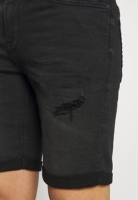 Shorts de mezclilla negra con un agujero deshilachado en el muslo delantero, dobladillos enrollados y una textura suave. Diseño casual con costuras tradicionales.