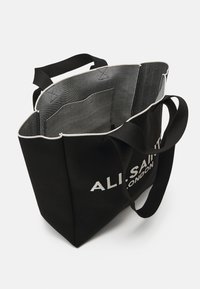 Sac tote noir avec doubles poignées, intérieur spacieux, une poche intérieure et texte blanc partiellement lisible "ALI SAINT LONDON" à l'extérieur.