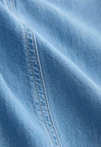 Detailný záber svetlomodrého rifľového materiálu zobrazujúci detailné prešívanie pozdĺž švu s viditeľnou textúrou a vzorom tkania.