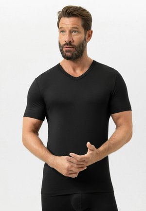 Bärtiger Mann mit kurzen Haaren, der ein eng anliegendes schwarzes V-Ausschnitt-T-Shirt trägt, leicht zur Seite schaut und die Hände vor sich verschränkt hält.