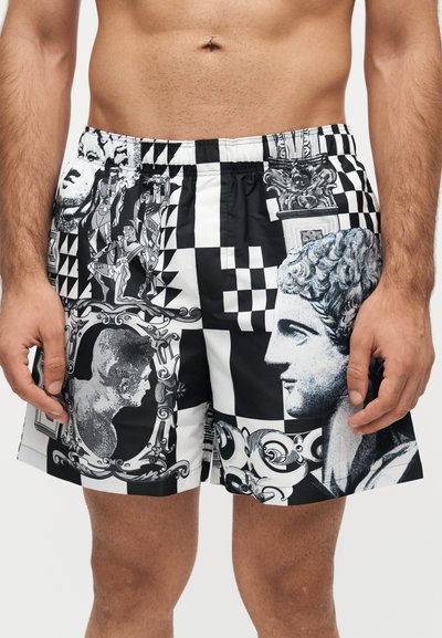 Shorts à motifs noir et blanc avec des imprimés artistiques de sculptures classiques, de visages et de dessins ornés portés par un mannequin masculin torse nu.
