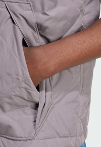 Ljusgrå quiltad jacka som visar en sidficka med en hand insatt. Tyget ser slätt ut med diamantsticking detaljer.