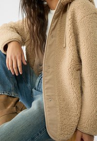 Beżowa kurtka z polaru o miękkiej, teksturowanej powierzchni, z beżową kontrastującą podszewką, zatrzaskami i kapturem. W połączeniu z niebieskimi dżinsami.