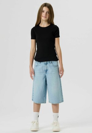 Persona giovane in piedi che indossa una maglietta nera a maniche corte, shorts di jeans larghi azzurri fino al ginocchio, calzini bianchi e scarpe da ginnastica bianche.