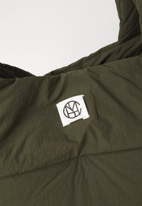Veste matelassée vert olive en tissu léger et texturé. Comprend un patch logo blanc brodé sur le devant et des détails de coutures visibles.