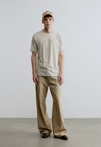 Bézs póló dombornyomott "BOSS" logóval, széles bézs nadrággal és hozzáillő sapkával párosítva. Sima anyag és laza szabás az egész ruhadarabon.