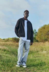 Veste zippée marron foncé portée sur un T-shirt blanc, associée à un jean large gris clair et des baskets noires et blanches. Fond de champ d'herbe.