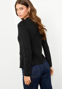 Pulover negru tricotat, cu mâneci lungi și guler rotund, având un detaliu subtil de peplum în spate și un buzunar lateral.