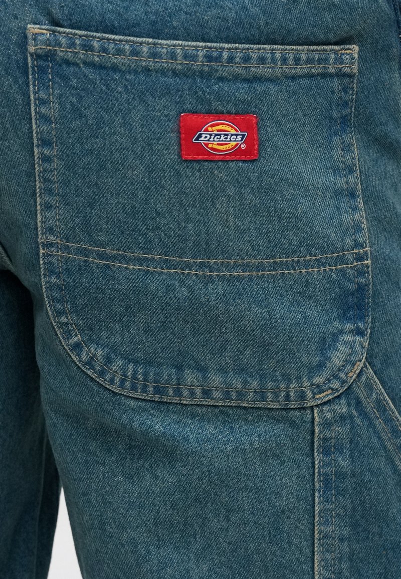 Pantaloni da lavoro in denim blu sbiadito con una grande tasca posteriore angolata. Presentano una toppa con il logo Dickies rossa e gialla sulla tasca superiore sinistra.