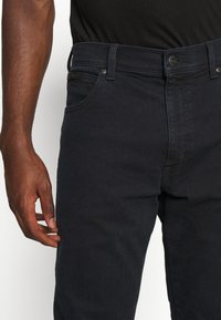 Jeans de denim oscuro con una textura suave, que presentan un diseño clásico de cinco bolsillos, un cierre de botón metálico y una sutil etiqueta de marca en la cintura.
