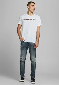 Weißes Baumwoll-T-Shirt mit schwarzem "JACK & JONES"-Logo, kombiniert mit dunkelblauen, distressed Jeans und schwarzen Sneakers, vor einem neutralen Hintergrund stehend.