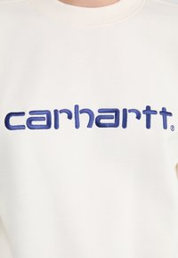 Hvid sweatshirt med blåt broderet "carhartt" logo. Stoffet er glat, med en standard rund halsudskæring og ribbede manchetter.