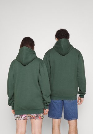 To personer står med ryggen mod hinanden iført matchende mørkegrønne hættetrøjer; den ene i blomster nederdel, den anden i blå denimshorts.