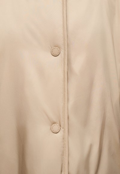 Κοντινό πλάνο ρούχου από μπεζ δέρμα με εμφανή ραφή και στρογγυλά μπεζ κουμπιά με ανάγλυφο το λόγκο «WEEKEND».