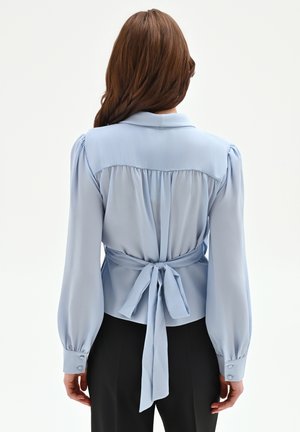 Donna con capelli castano lunghi che indossa una camicetta azzurra con maniche a sbuffo e un fiocco annodato in vita, abbinata a pantaloni neri, vista da dietro.