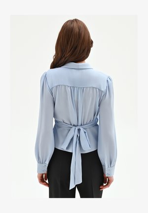 Donna con capelli castano lunghi che indossa una camicetta azzurra con maniche a sbuffo e un fiocco annodato in vita, abbinata a pantaloni neri, vista da dietro.