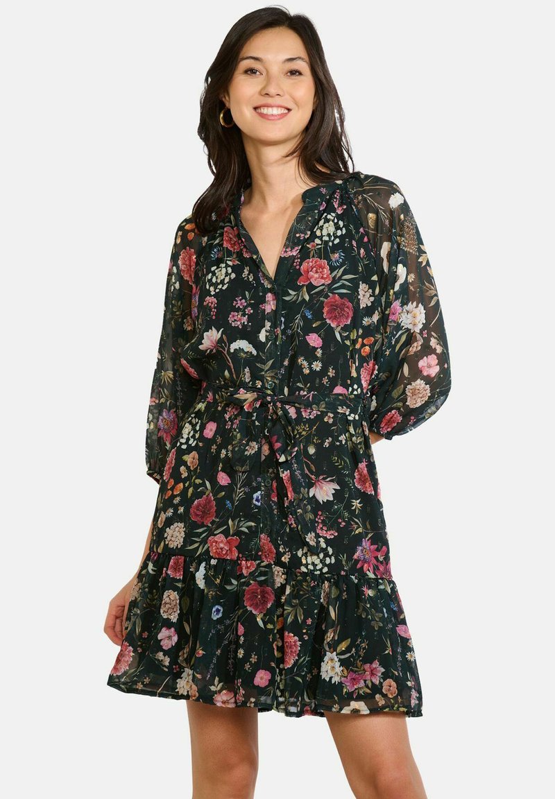 Kobieta z ciemnymi włosami, ubrana w czarną sukienkę w kwiaty z przezroczystymi długimi rękawami, wiązaniem w talii i falbaniastym dołem, uśmiechająca się na białym tle.