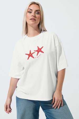 Kvinde med glat blondt hår iført en hvid oversized T-shirt med to røde søstjerner og tekst, kombineret med blå jeans foran en ensfarvet baggrund.