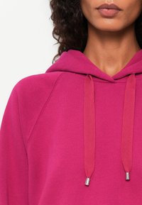 Fuchsia Kapuzenpullover mit weichem Stoff, ausgestattet mit einer Kordelzugkapuze und silberfarbenen Spitzen an den Schnürsenkeln. Das Design umfasst Raglanärmel.